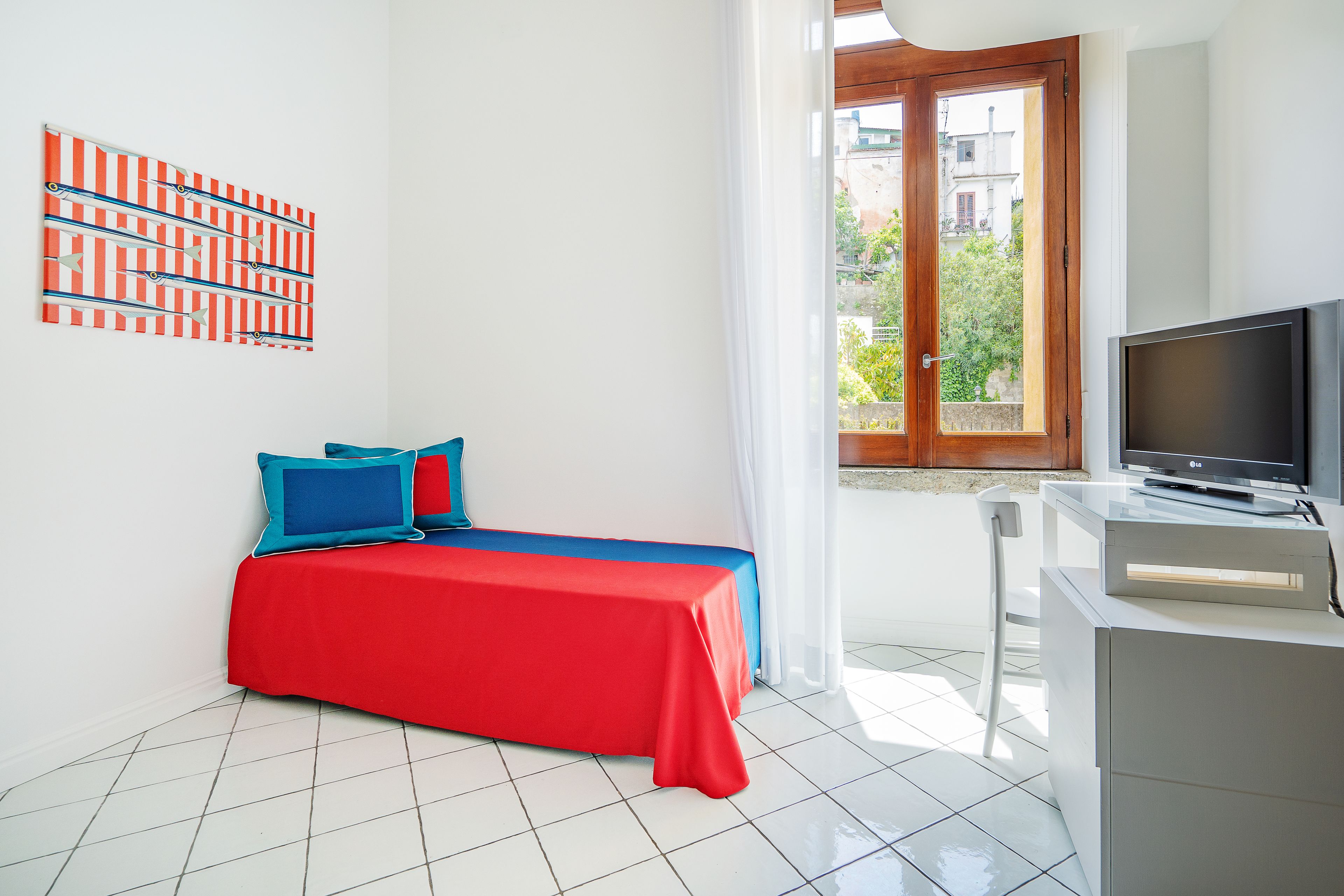 Un dormitorio con una colcha roja, un televisor sobre un mueble bajo y una ventana con vistas a edificios.