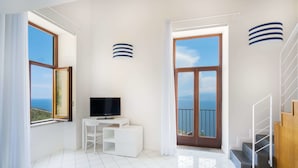 Um quarto claro com televisão, escrivaninha e vista para o mar.
