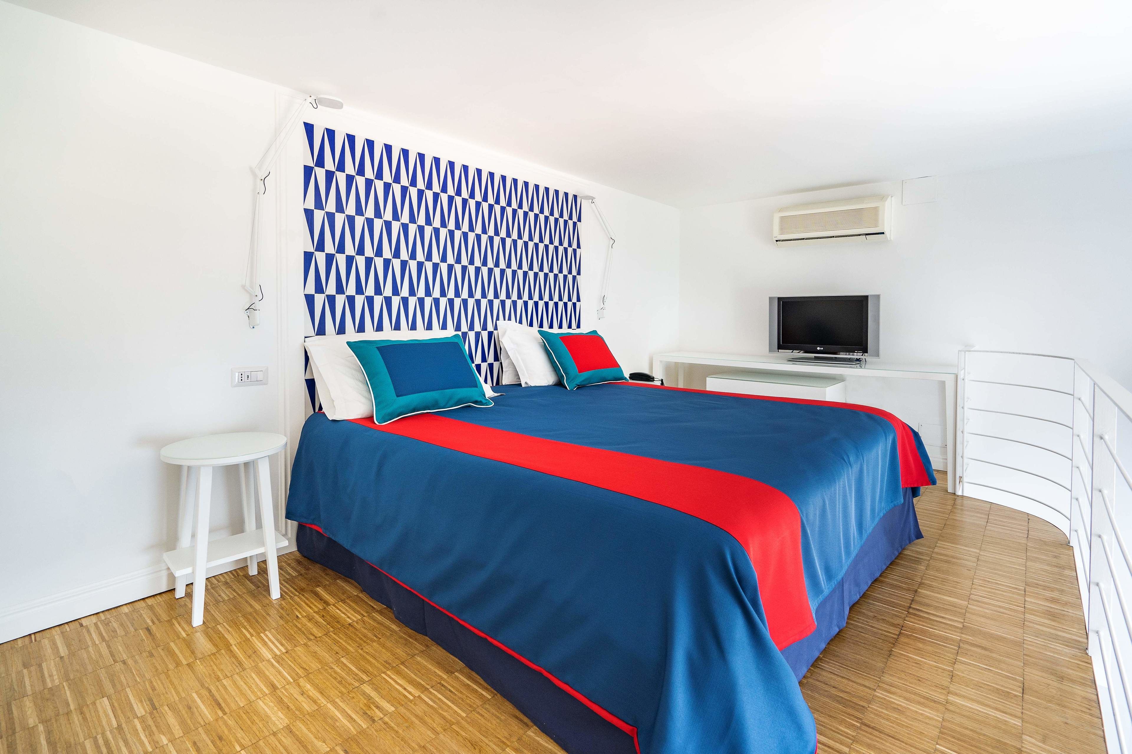 Un dormitorio con cama, televisión, mesita auxiliar blanca y cabecera estampada.