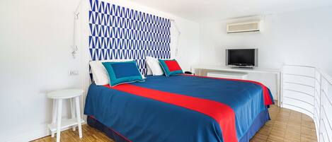 Un dormitorio con cama, televisión, mesita auxiliar blanca y cabecera estampada.