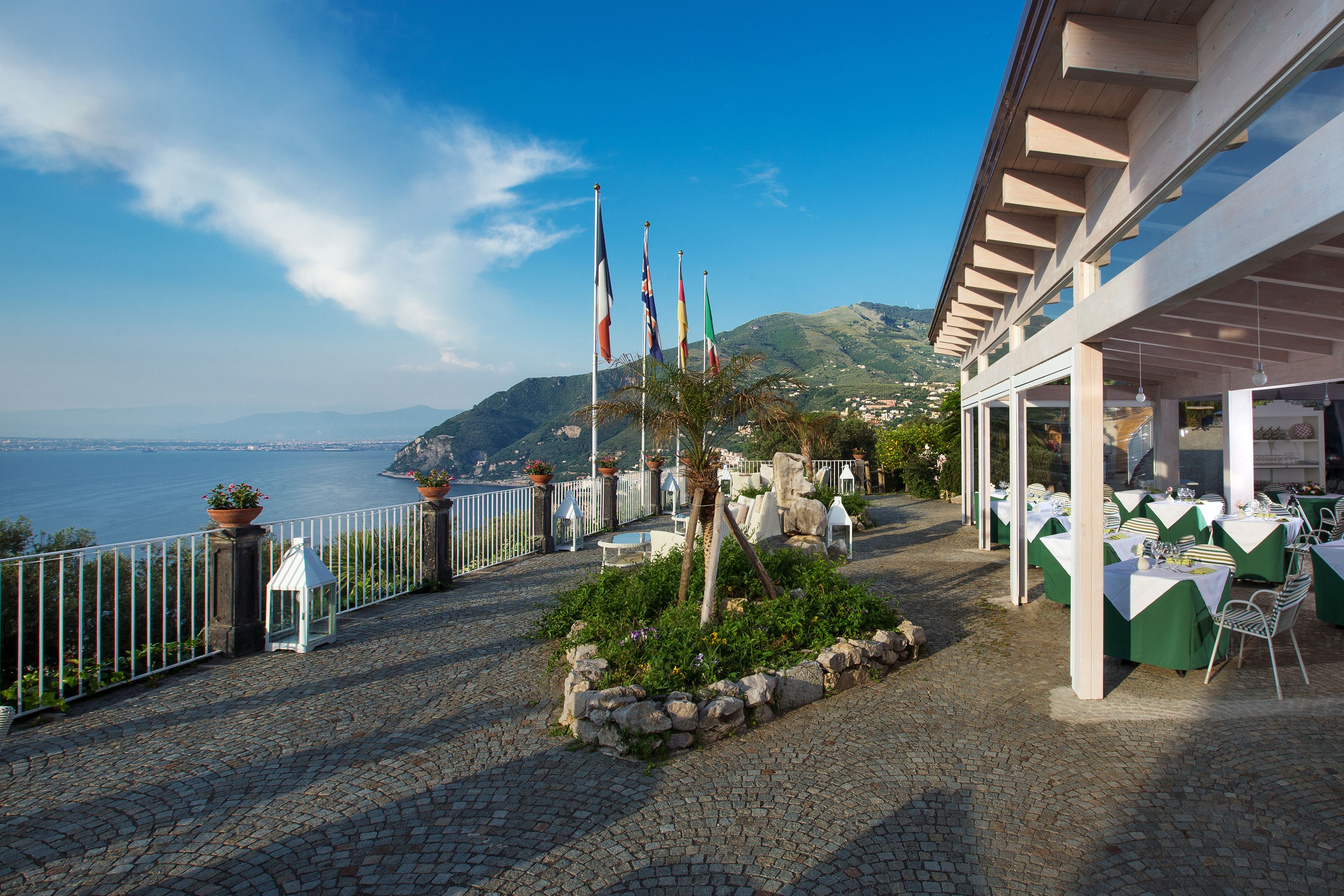 A terrace with tables set for dining, overlooking a coastal view with flags and greenery.