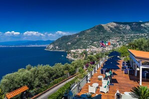 Eine Seeterrasse mit Sitzgelegenheiten im Freien, mit Blick auf eine Küstenstadt und das Meer.