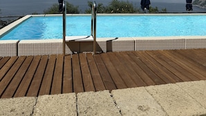 Um deck de madeira com piscina, duas pessoas vestidas de escuro e vista para o mar e montanhas.