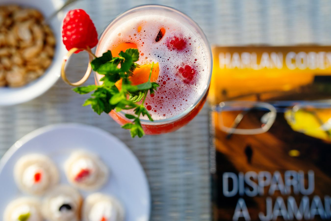 A glass of beverage with a strawberry garnish, a book titled "DISPARU A JAMAIA" and a plate of food in the background.