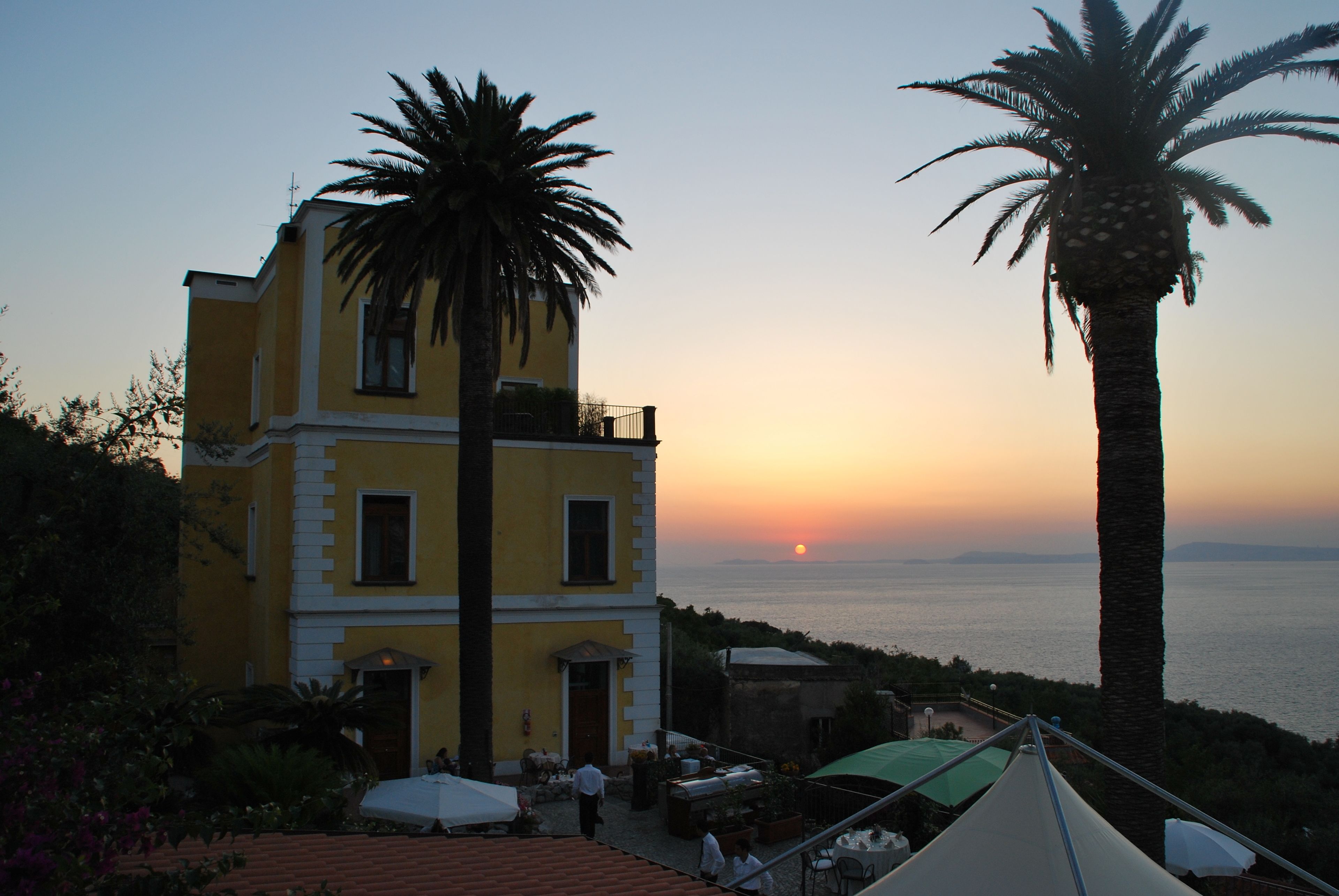 Un edificio giallo con un balcone che si affaccia sul mare, palme e posti a sedere all'aperto.