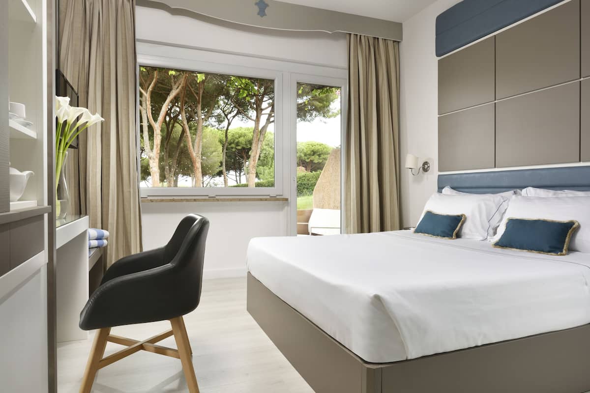 Un dormitorio con cama, una silla, una ventana con cortinas y vistas a árboles.