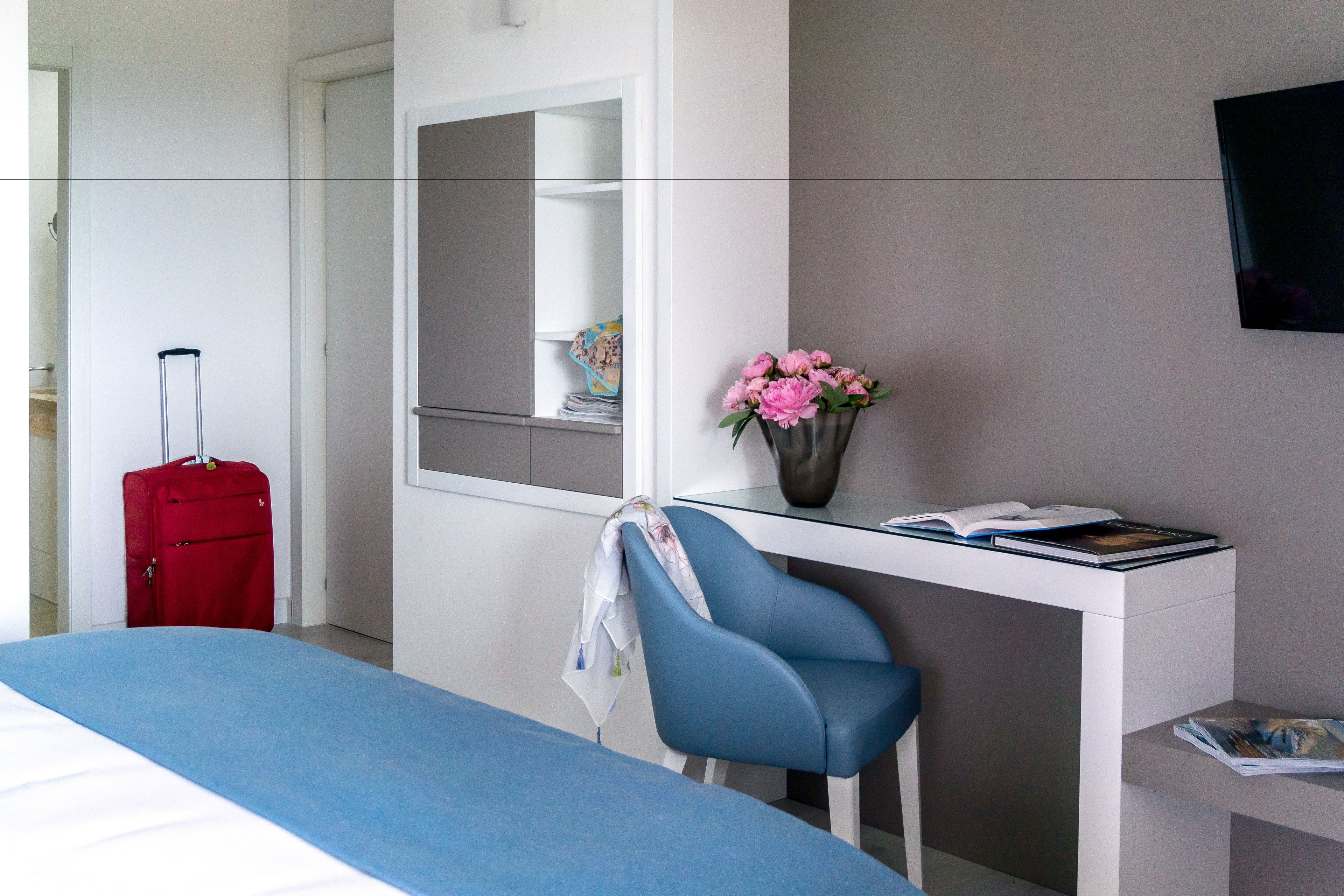 Ein Hotelzimmer mit einem Bett, einem Schreibtisch mit Stuhl, einem Spiegel, einer Vase mit Blumen und einem roten Koffer.