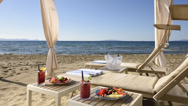 Mesa en la playa con una bandeja de frutas, bebidas y una silla de playa bajo una sombrilla.