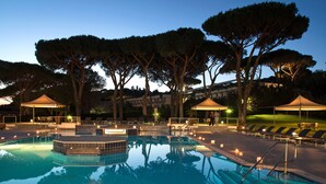 Una piscina con sillones y sombrillas, rodeada de árboles y un edificio al fondo.