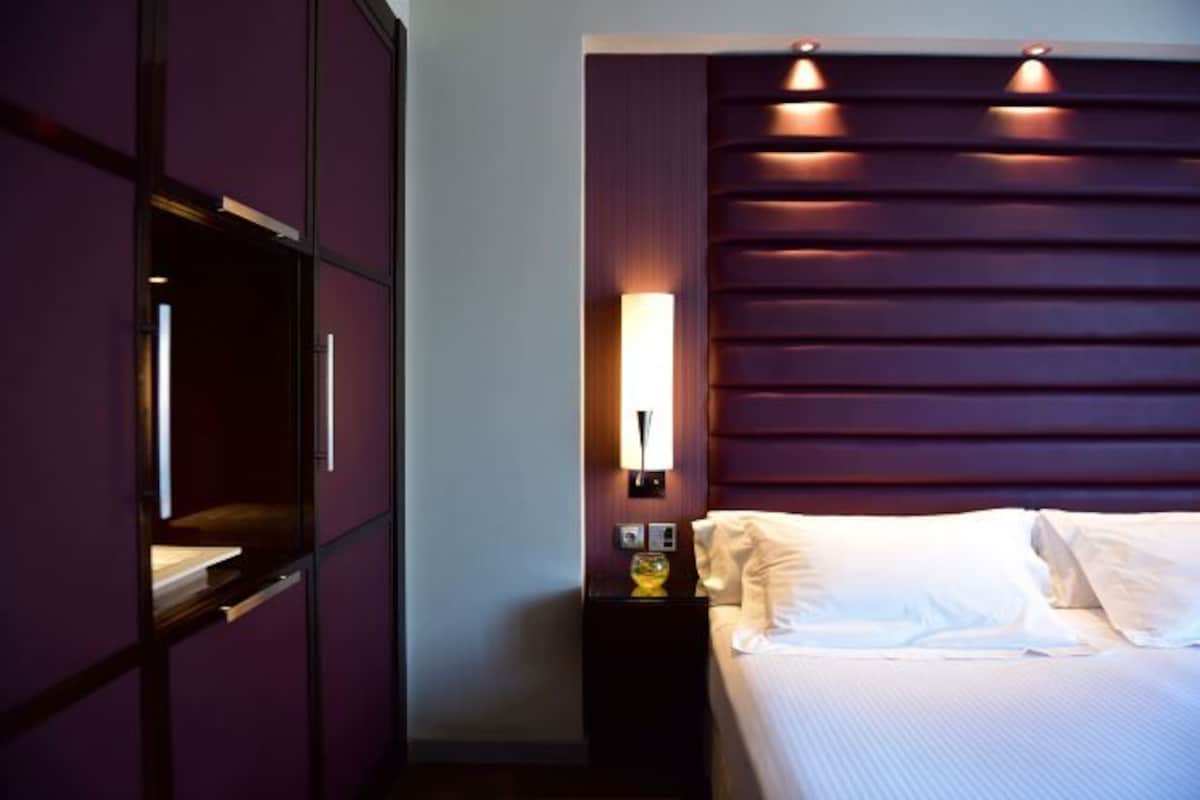 A hotel room with a bed, a nightstand, a wall-mounted TV, and a wall-mounted light fixture.