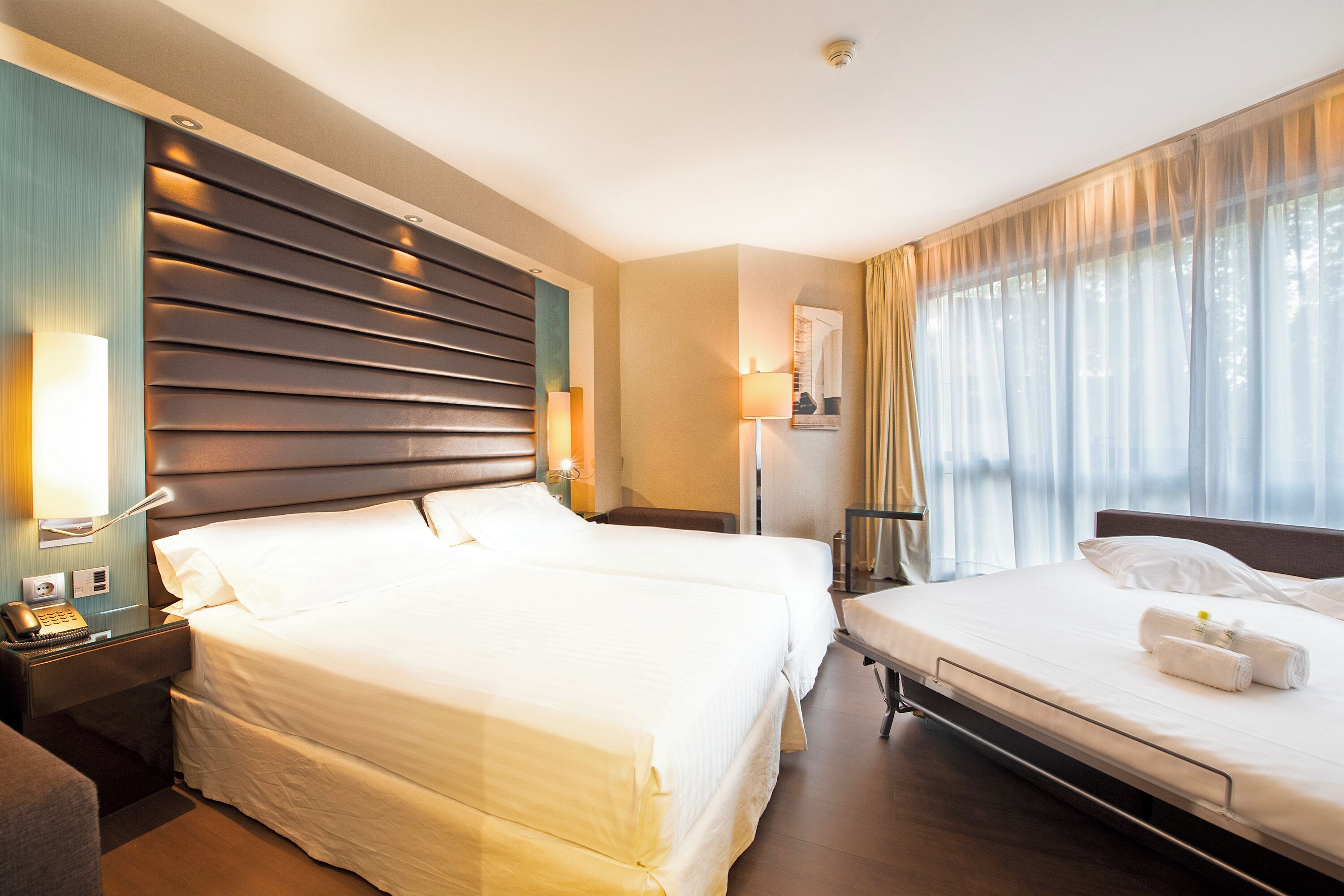 A hotel room with two beds, a nightstand with a lamp, a wall-mounted TV, and a window with sheer curtains.