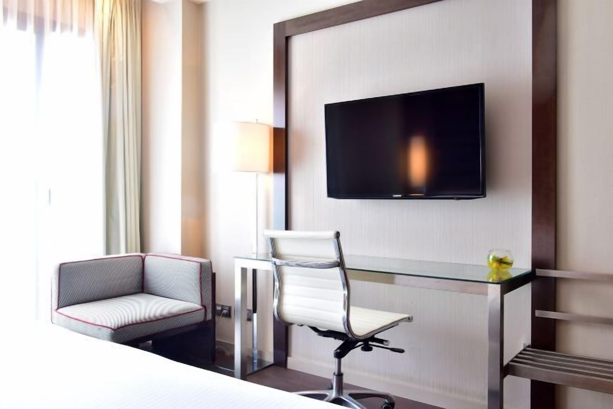 A hotel room with a bed, a desk with a chair, a television mounted on the wall, and a window with curtains.