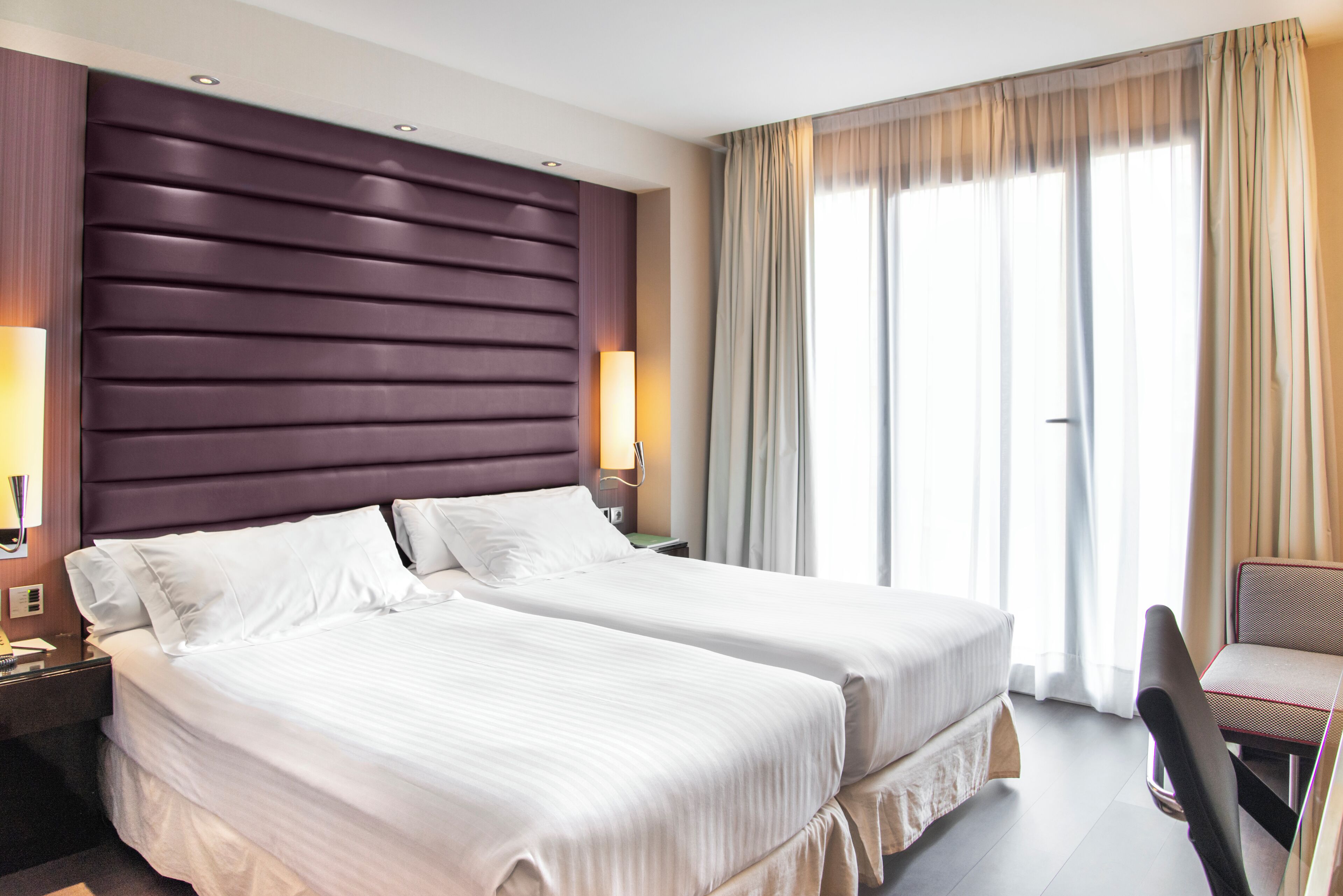 A hotel room with two beds, a large padded headboard, two bedside lamps, a chair, and a large window with curtains.