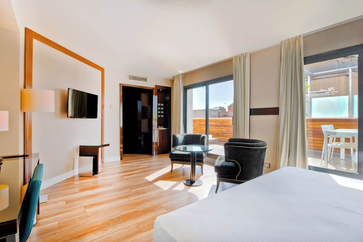 Habitación de hotel con cama, escritorio, silla, televisor y balcón con vistas.