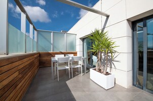 Un espacio al aire libre moderno con sillas blancas, un banco de madera, una planta en maceta y un muro de cristal.