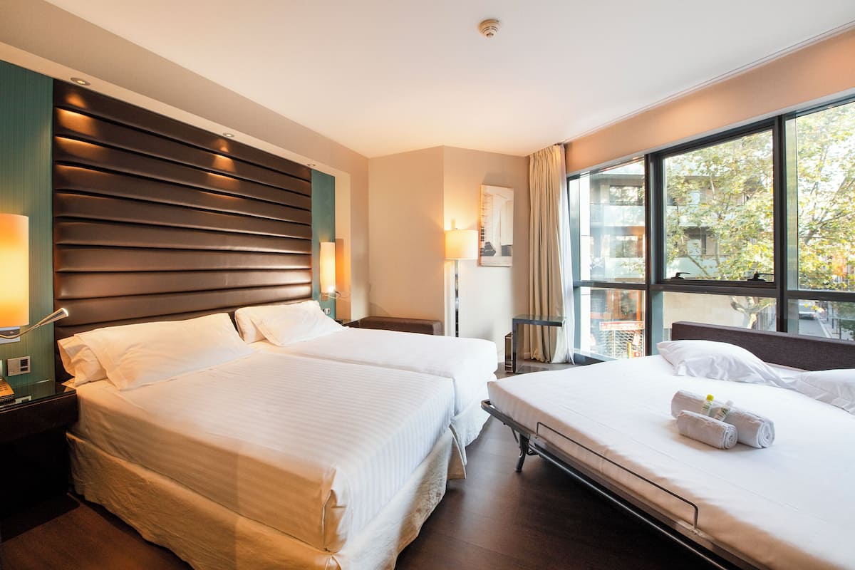 Habitación de hotel con dos camas, un ventanal grande y un cabecero moderno.