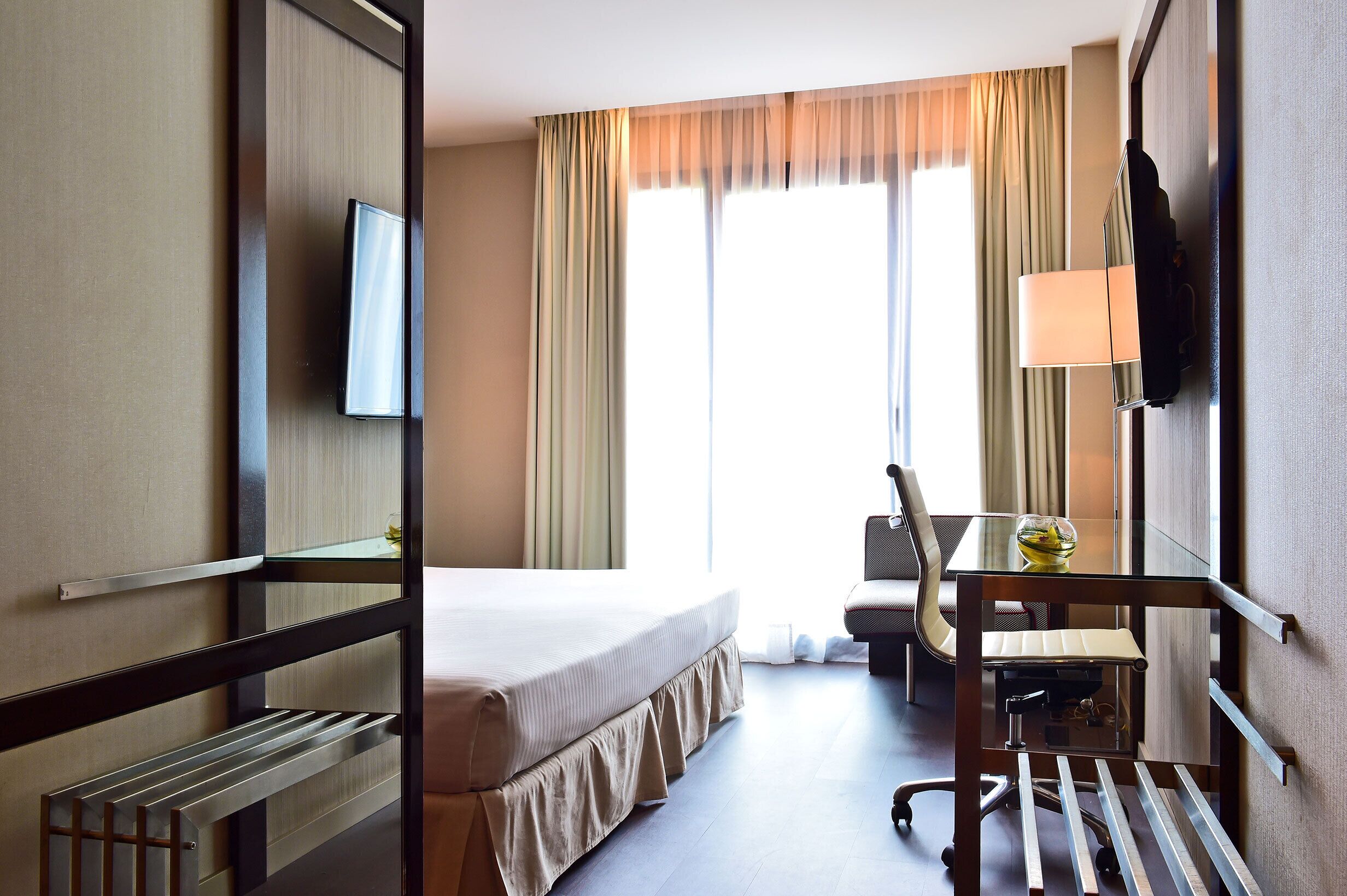 Habitación de hotel con cama, escritorio y silla, televisión y ventana con cortinas.