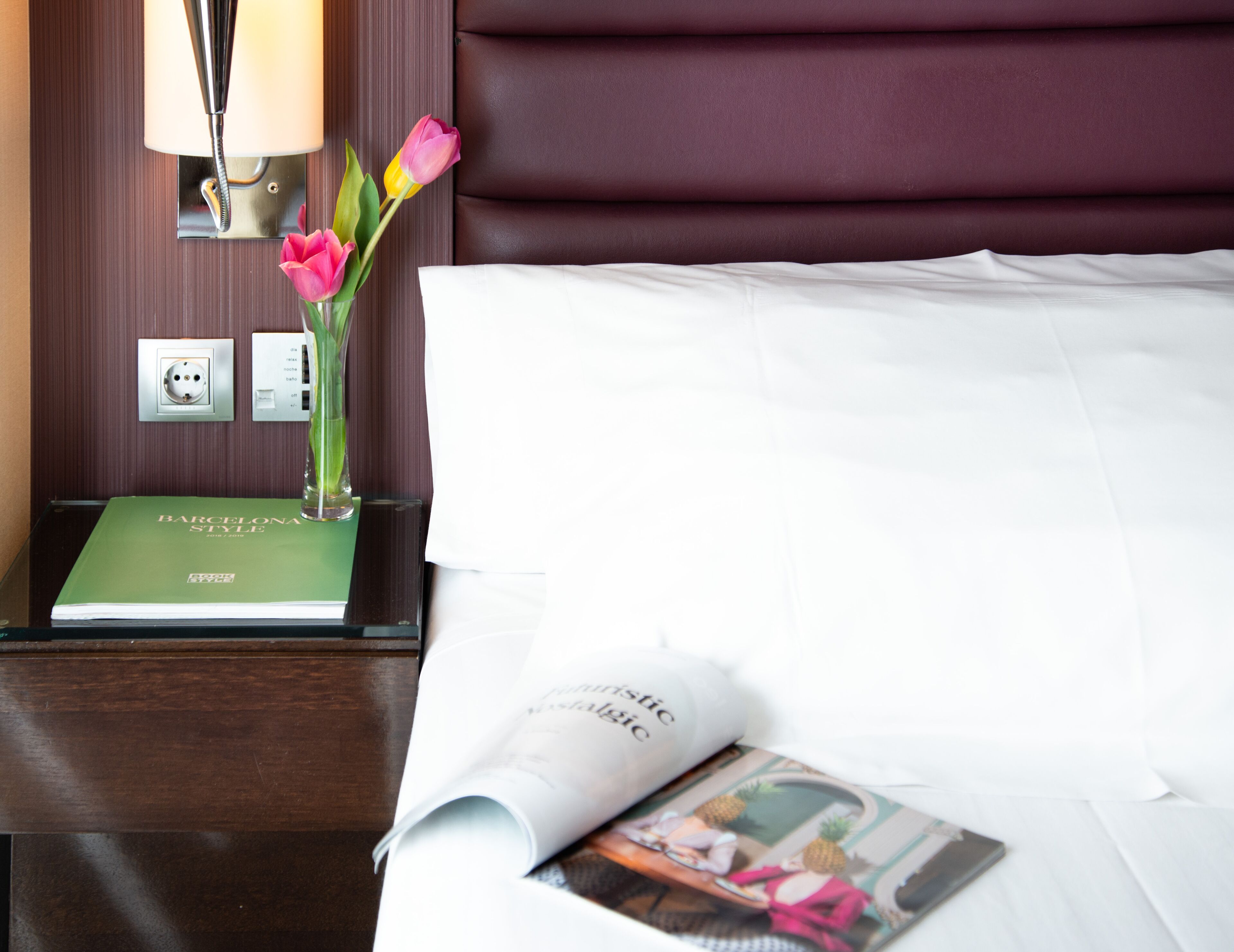 A neatly made bed with white linens, a green book titled 'BARCELONA STYLE', a lamp, and a vase with pink flowers on a wooden nightstand.