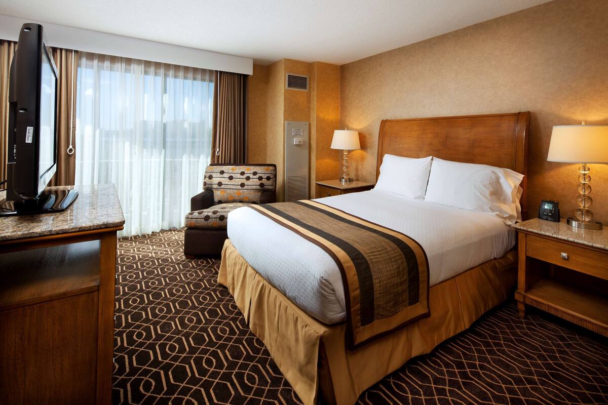A hotel room with a large bed, a desk with a television, a chair, and a nightstand with a lamp.