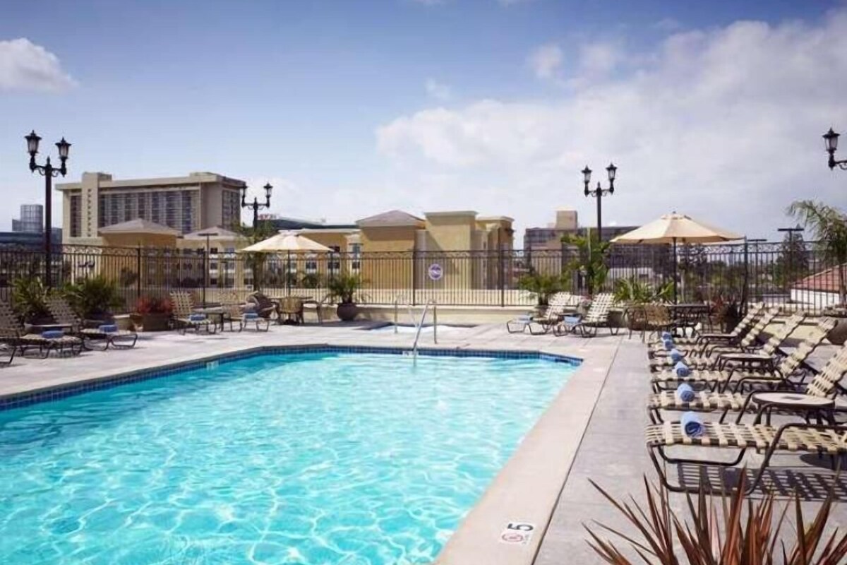 A swimming pool with lounge chairs and umbrellas, surrounded by a fence and buildings in the background.