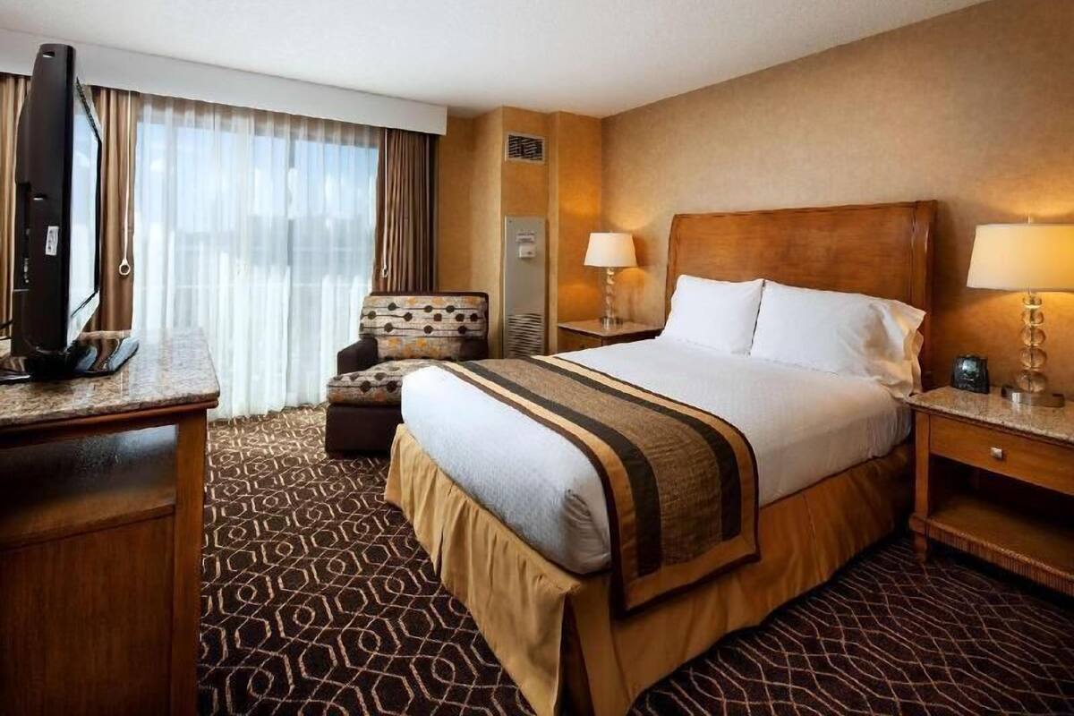 A hotel room with a large bed, a desk with a television, a chair, a bedside table with a lamp, and a window with curtains.