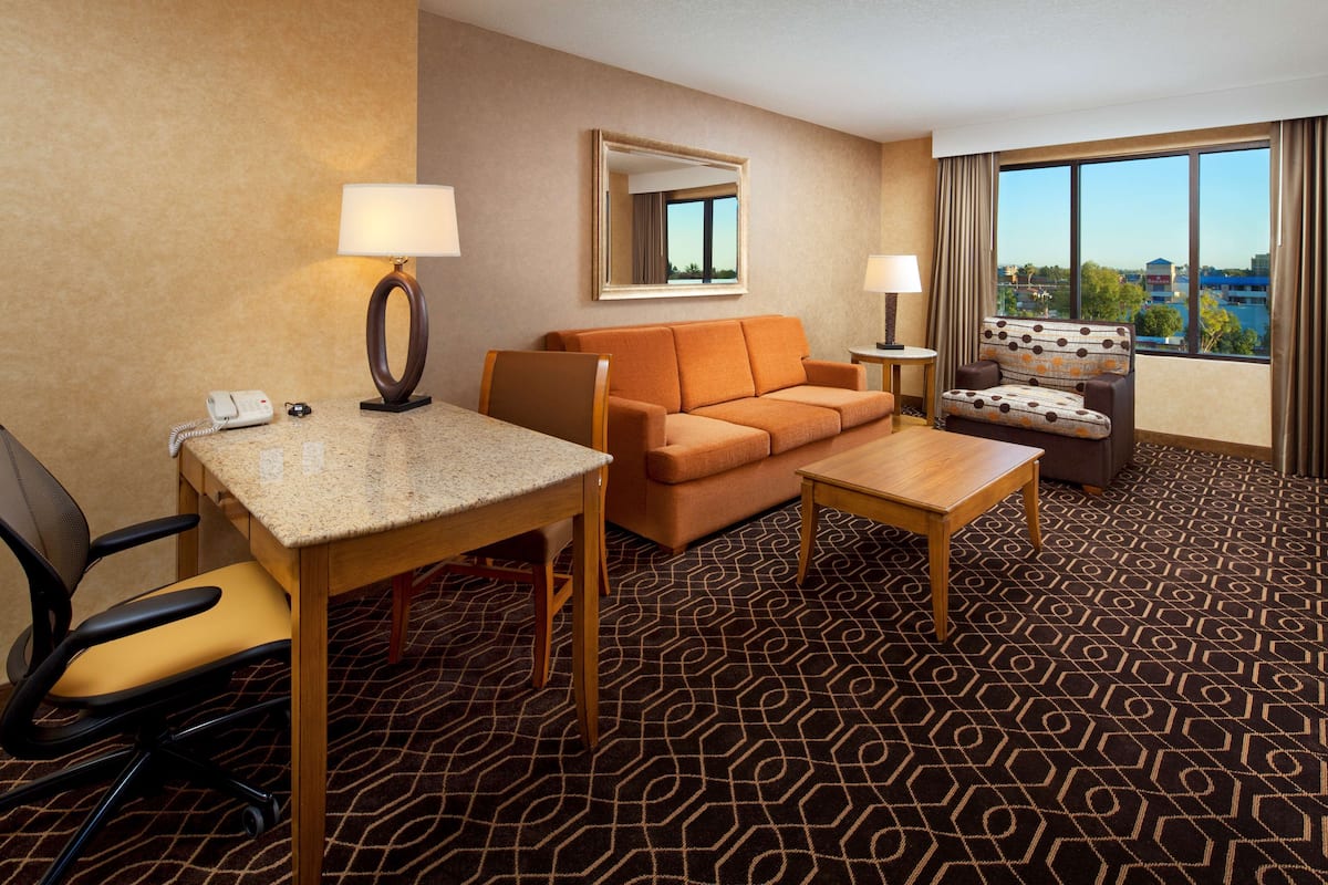 A hotel room with a sofa, a dining table, a chair, a lamp, a mirror, and a view of the city.