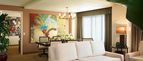 Habitación de hotel con un sofá color beige, una mesa de comedor con sillas, un cuadro grande en la pared y una planta en maceta.