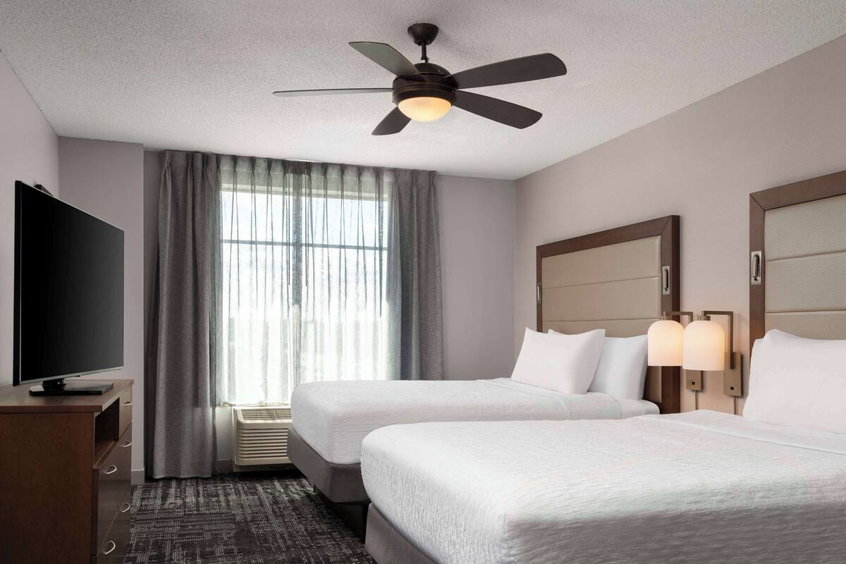 Une chambre d’hôtel avec deux lits, un téléviseur à écran plat, un ventilateur de plafond et une fenêtre avec des rideaux.