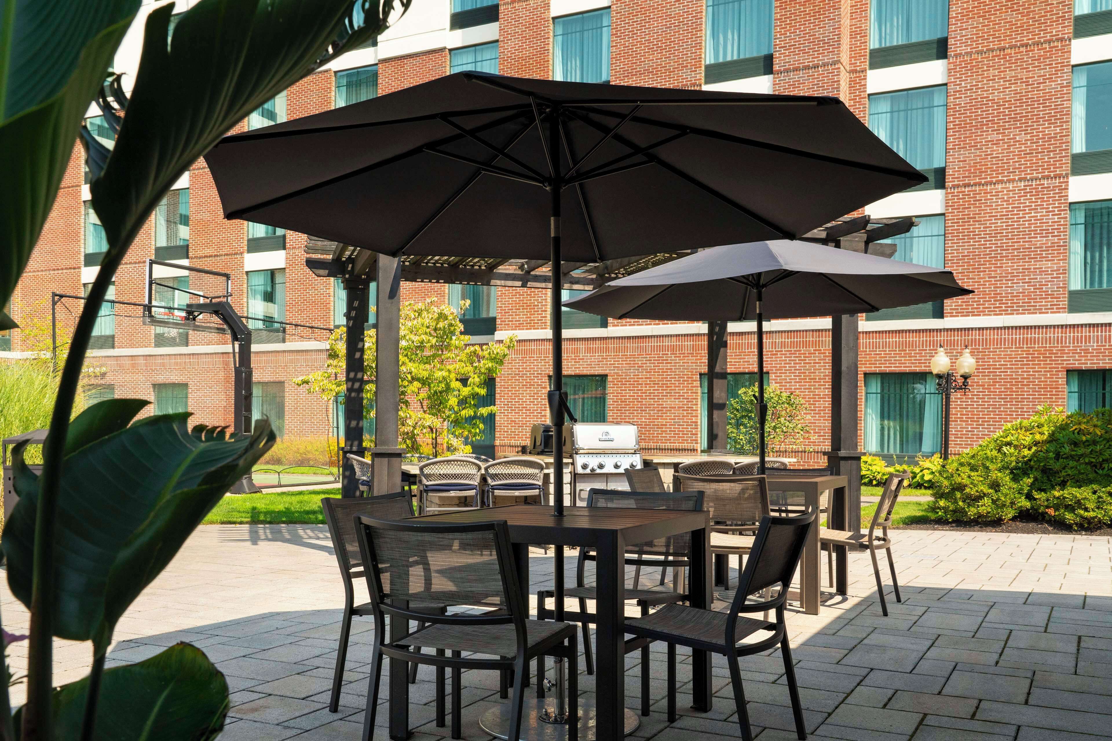 An outdoor patio with a table and chairs, a grill, and a brick building in the background.