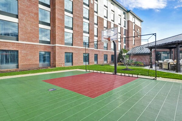 A basketball court with a red and green surface, surrounded by a brick building with large windows and a covered seating area.