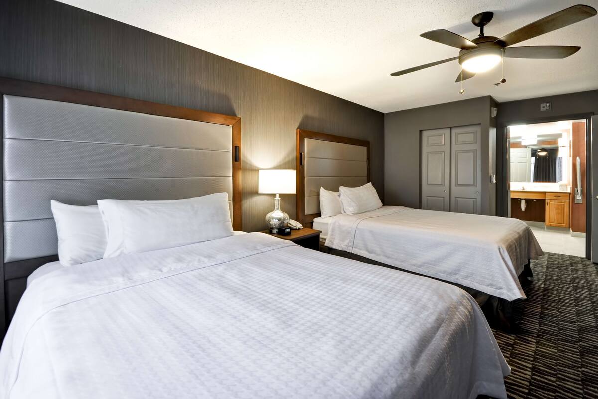 A hotel room with two beds, a ceiling fan, a nightstand with a lamp, and a mirror.