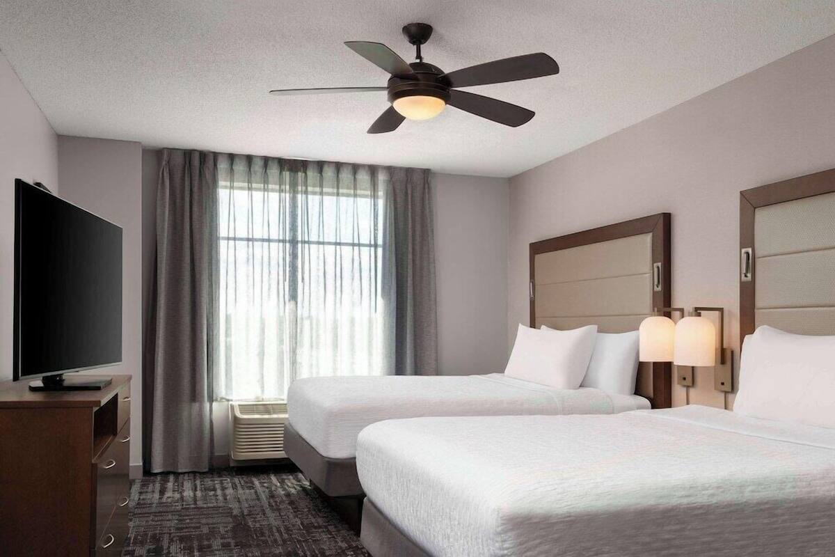 A hotel room with two beds, a ceiling fan, a TV, a window with curtains, and wall-mounted lamps.