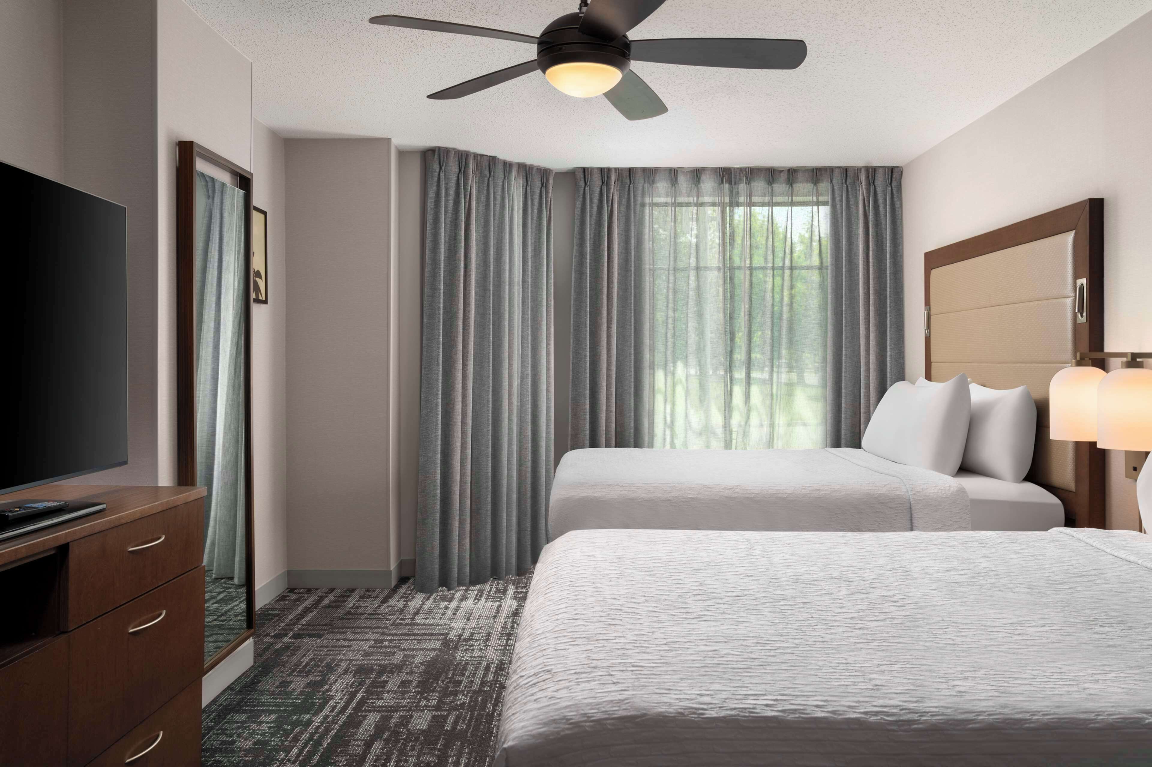 A hotel room with two beds, a ceiling fan, a television, and a large window with curtains.