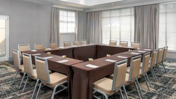 A conference room with a rectangular table surrounded by chairs, large windows with curtains, and a patterned carpet.