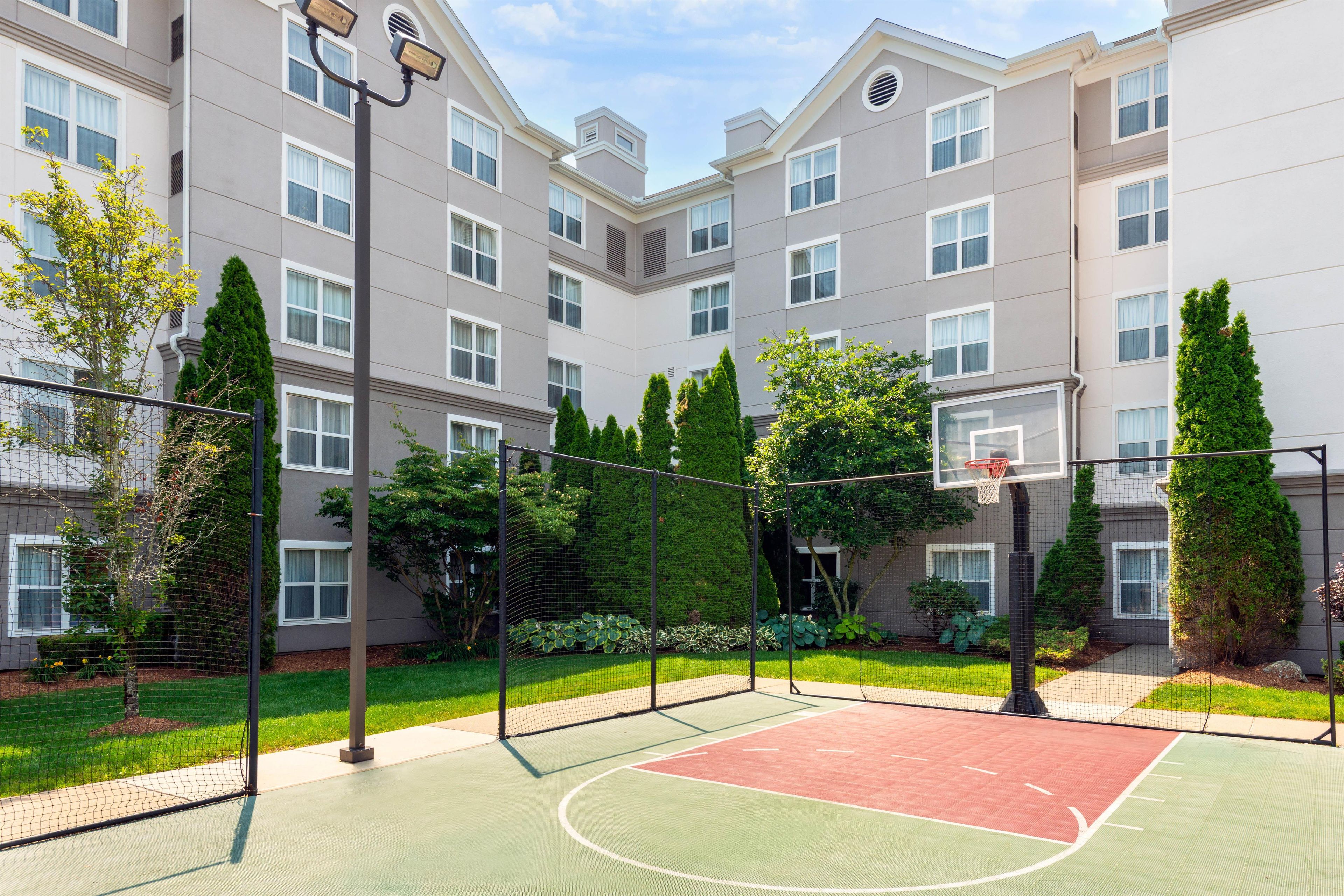 Uma quadra de basquete com rede, cercada por uma grade, e um edifício com várias janelas ao fundo.