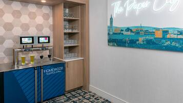 A hotel bar area with a neon sign reading 'The Paper City,' a selection of glasses on shelves, and a digital screen displaying beverage options.