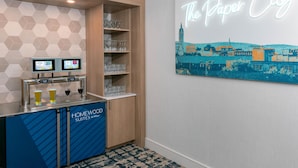 A hotel bar area with a neon sign reading 'The Paper City,' a selection of glasses on shelves, and a digital screen displaying beverage options. - Homewood Suites by Hilton Holyoke-Springfield/North (Holyoke)