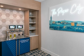 A hotel bar area with a neon sign reading 'The Paper City,' a selection of glasses on shelves, and a digital screen displaying beverage options. - Homewood Suites by Hilton Holyoke-Springfield/North (Holyoke)