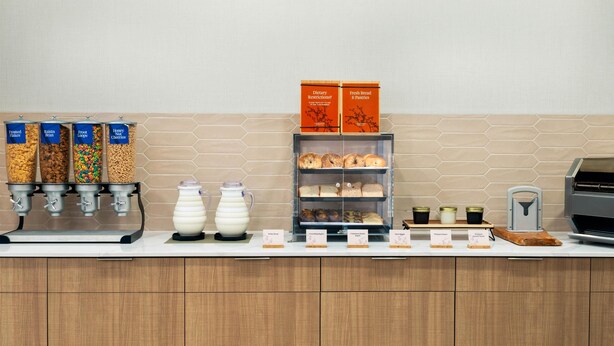 A breakfast bar with cereal dispensers, a coffee machine, milk pitchers, and a display of pastries. - Homewood Suites by Hilton Holyoke-Springfield/North (Holyoke)