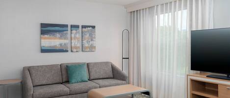A modern hotel room with a grey sofa, a small table, a flat-screen TV, and two abstract paintings on the wall.