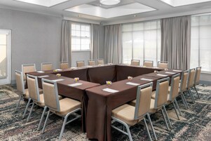 Una sala de conferencias con una mesa rectangular rodeada de sillas, ventanas amplias con cortinas y una alfombra estampada.