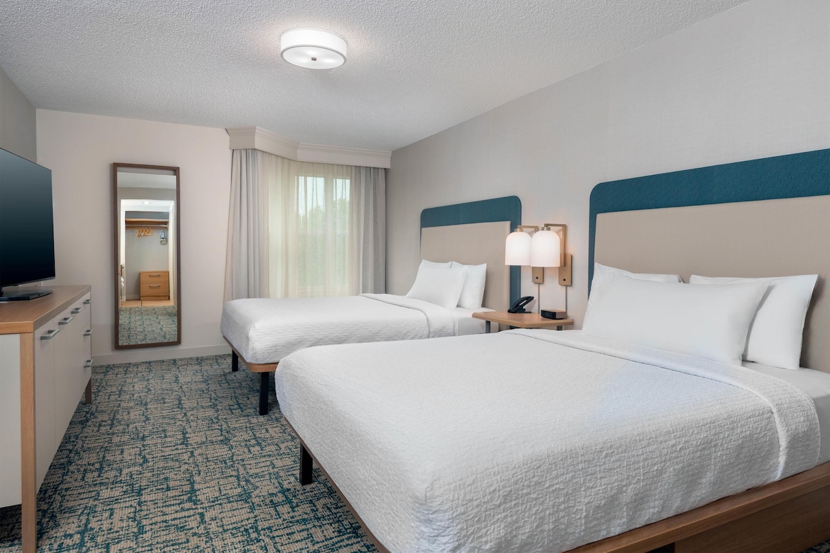 A hotel room with two beds, a desk with a television, a mirror, and a window with curtains.