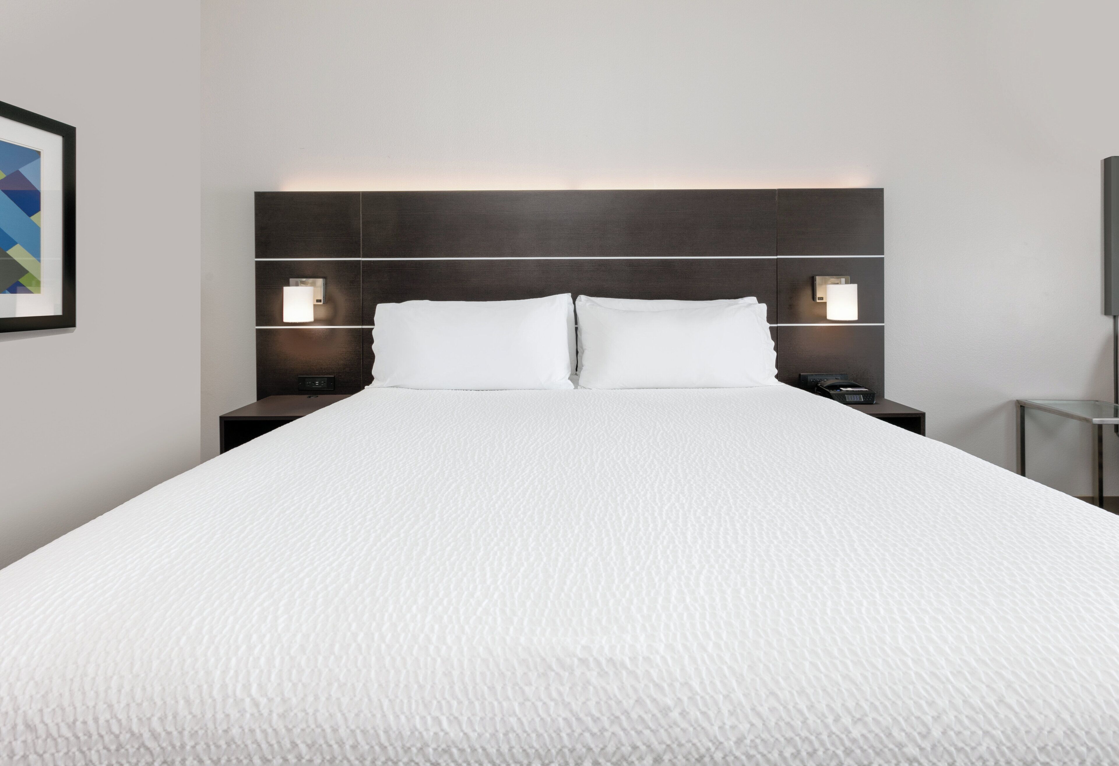 Una cama grande con cubre cama blanco texturizado, dos mesitas de noche con lámparas y un cabecero oscuro.