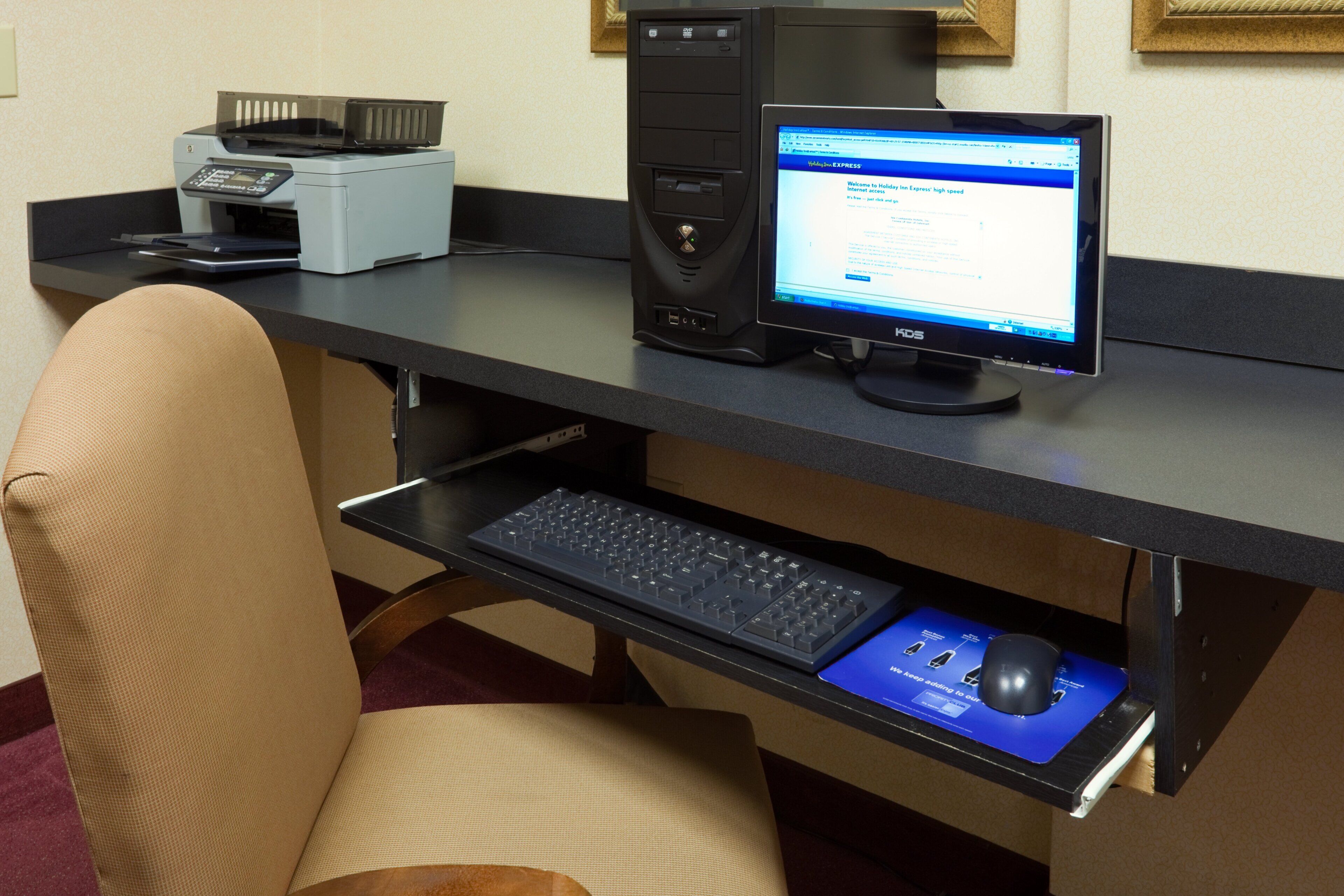Un espacio de trabajo en un hotel, con una computadora de escritorio, una impresora, un teclado y un ratón sobre un escritorio.