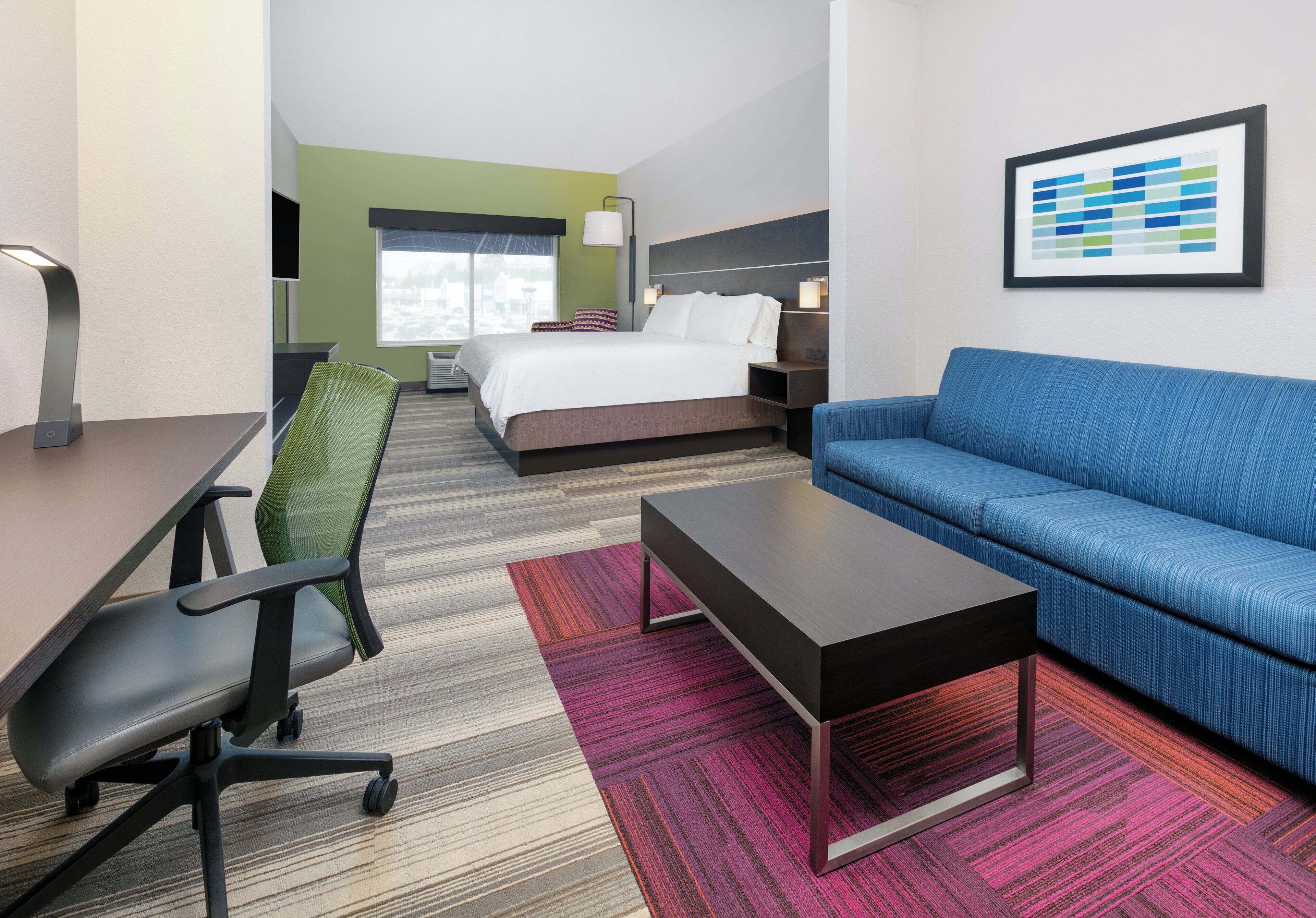 A hotel room with a bed, a blue sofa, a desk with a chair, and a colorful rug.