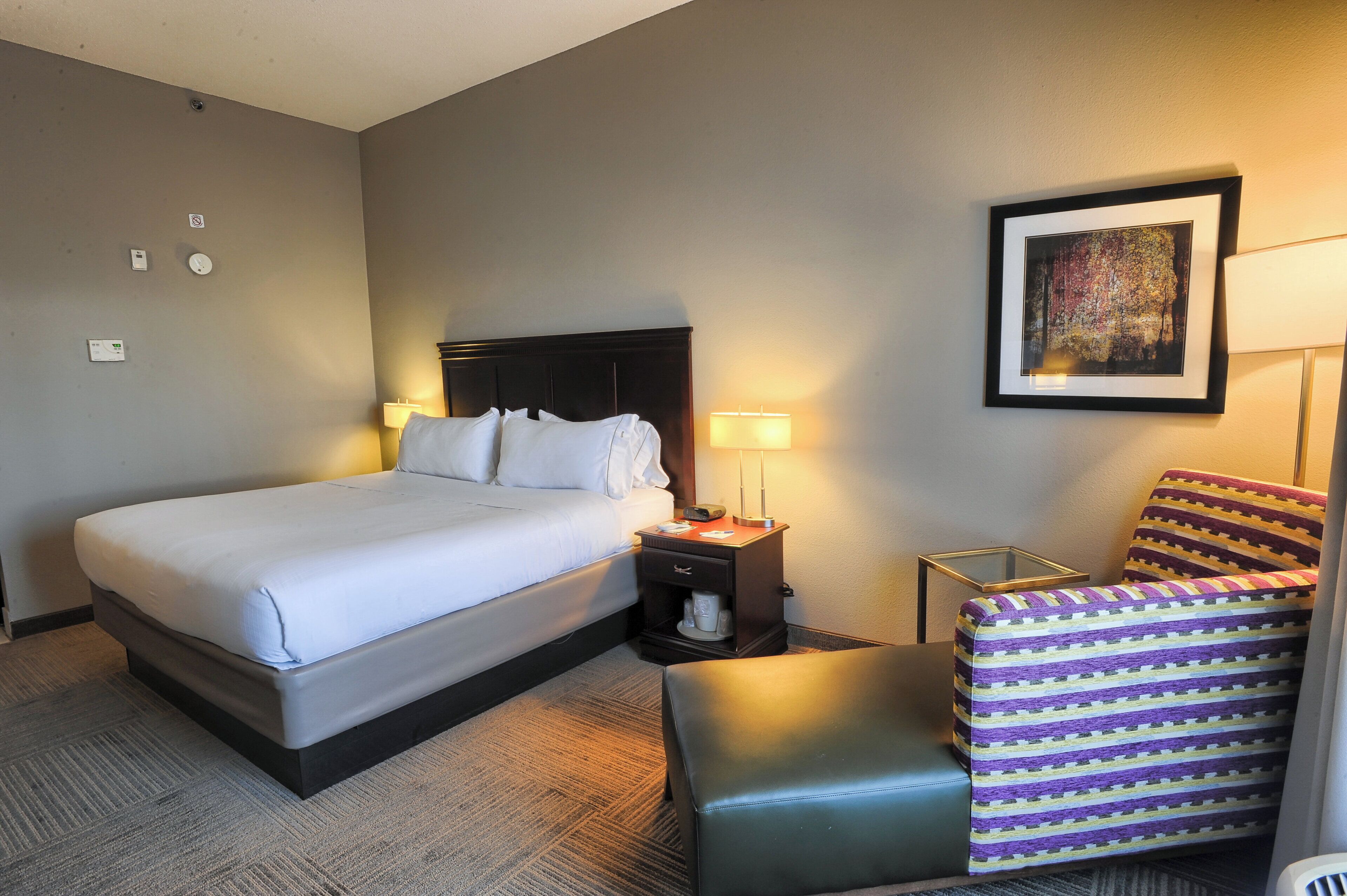 A hotel room with a bed, bedside lamps, a chair, a small table, and a painting on the wall.