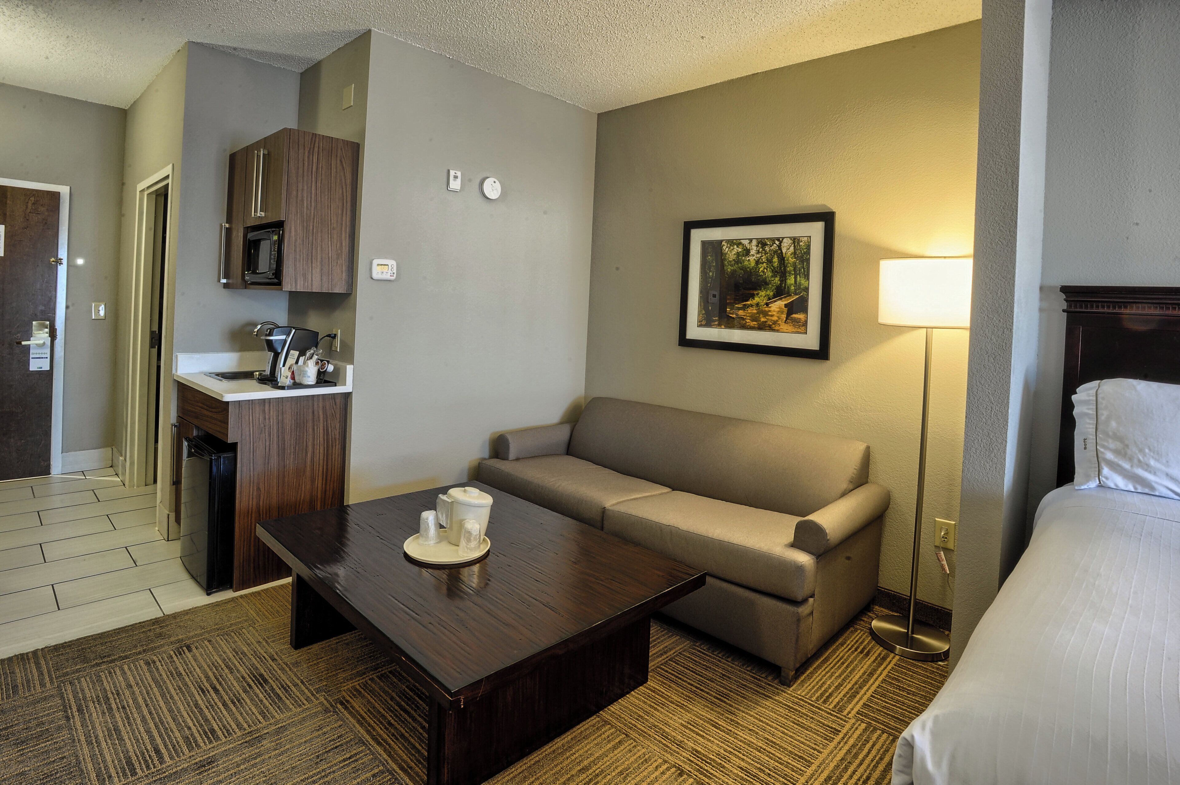 A hotel room with a kitchenette, a sofa, a coffee table, a bed, and a bedside lamp.