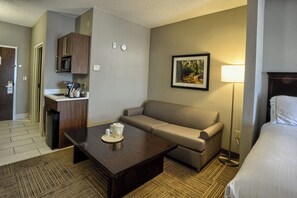 A hotel room with a kitchenette, a sofa, a coffee table, a bed, and a bedside lamp. - Holiday Inn Express Hotel & Suites Pell City by IHG (Pell City)