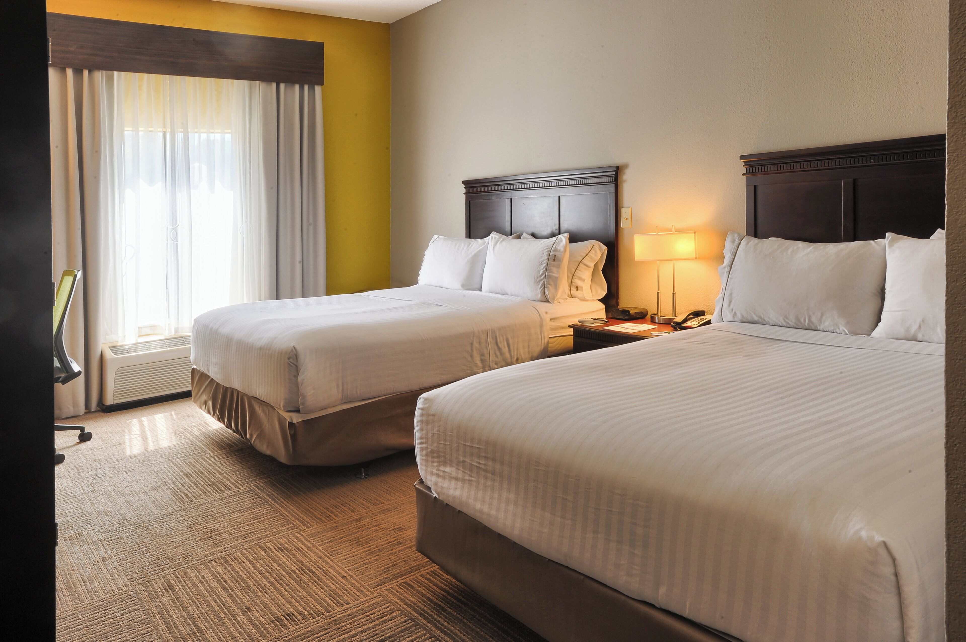 A hotel room with two beds, a nightstand, a lamp, and a window with curtains.