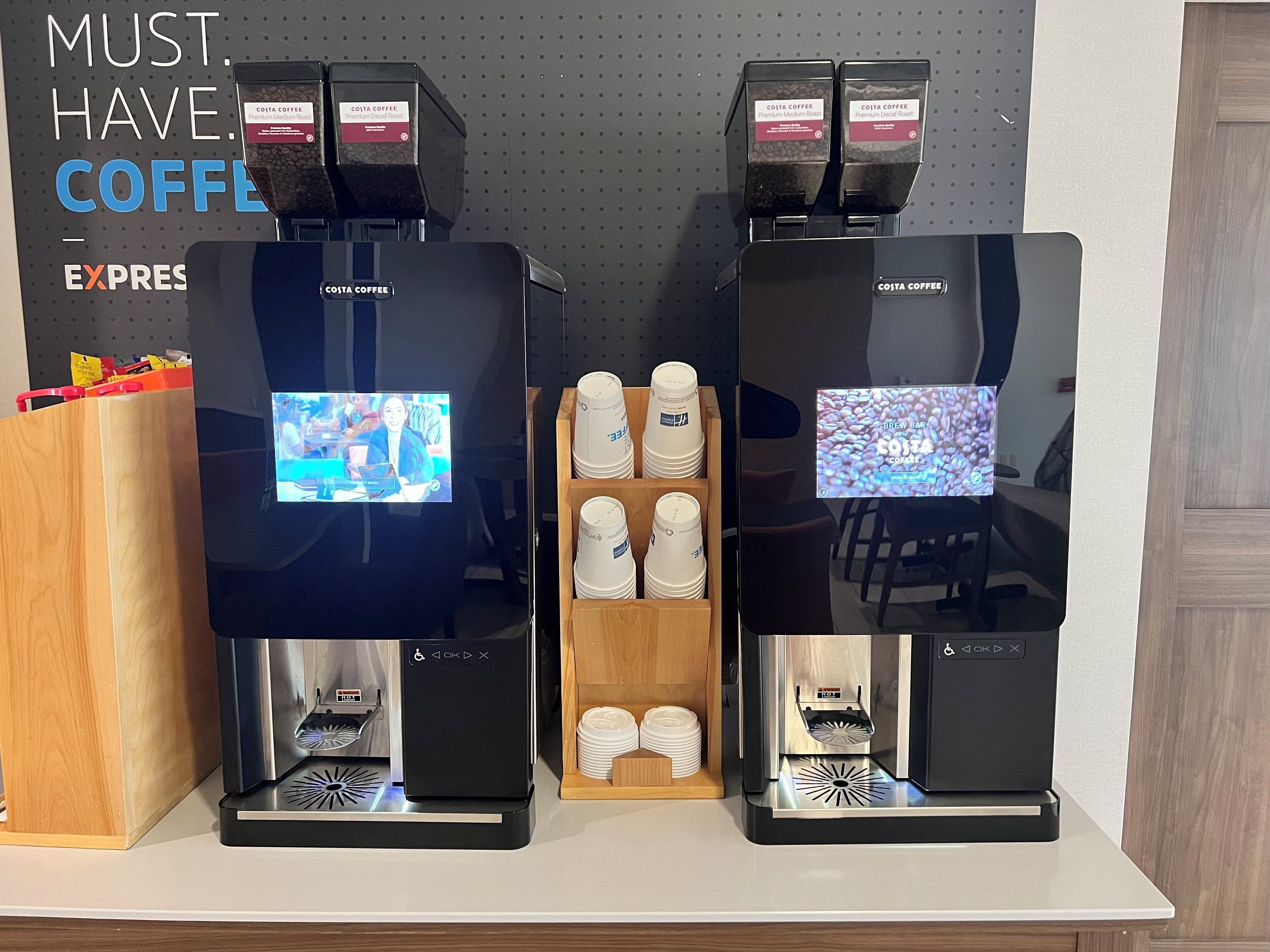 Een koffiehoek met twee espresso machines, een presentatie van wegwerpbekers en een scherm dat een koffievideo toont.